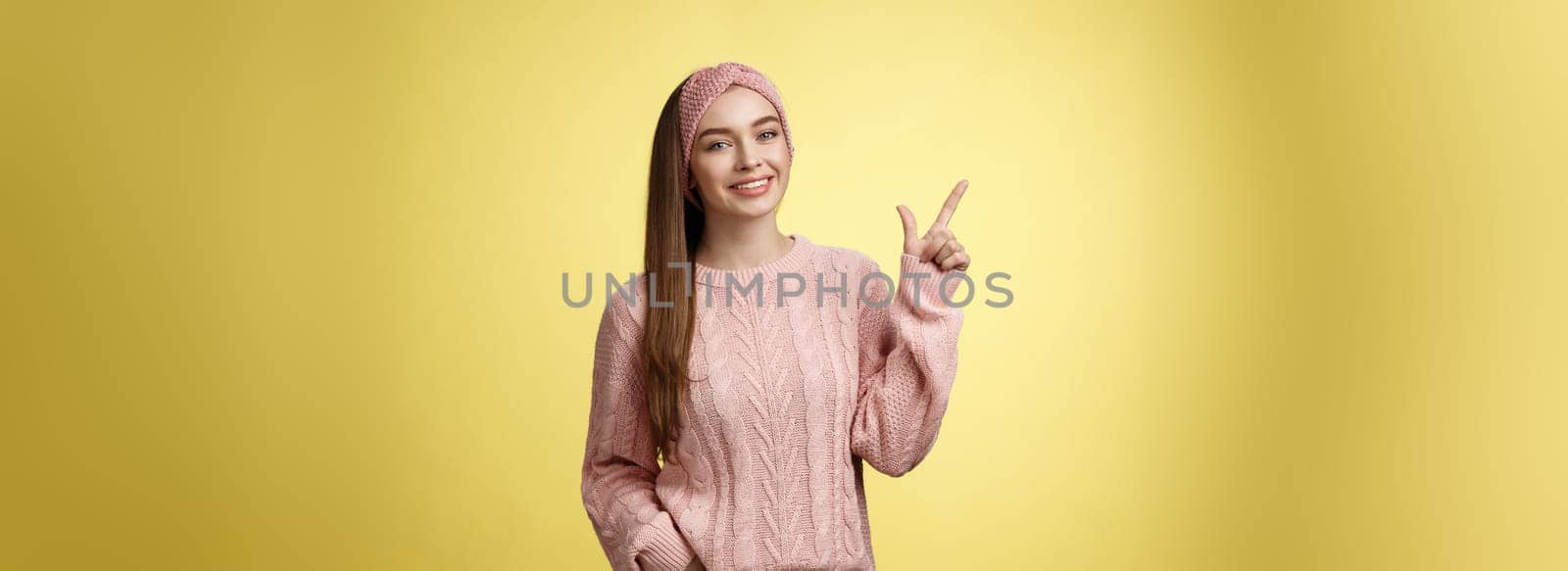 Girl picked staff, made decision pointing up at product smiling pleased. Studio shot of young student in knitted sweater, grinning indicating upper left corner enthusiastic, giving recommendation.