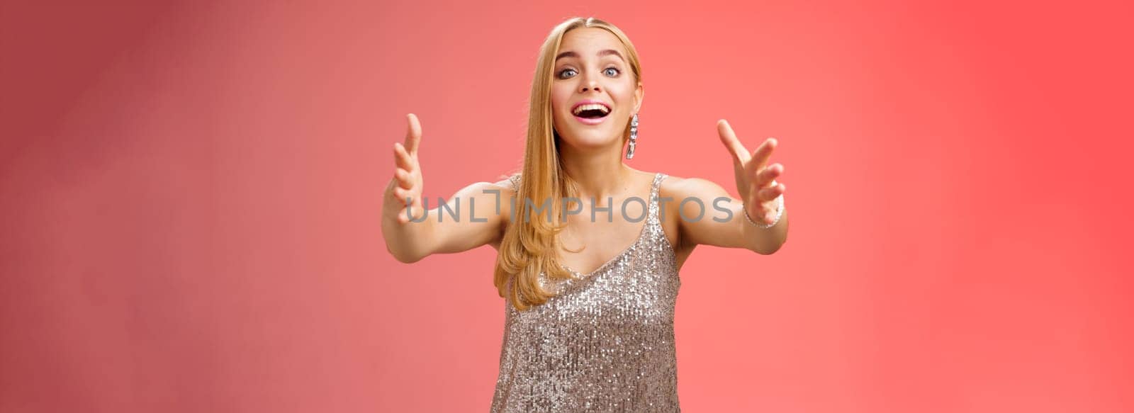 Excited charmign touched hearwarming young blond woman in silver party glamour dress stretch hands towards camera amused wanna hug cuddle hold cute puppy arms, standing red background.