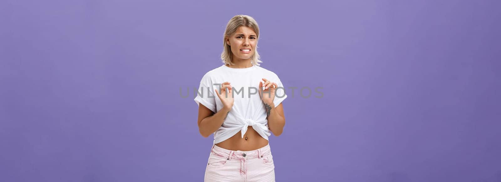 Ew take disgusting thing away from me. Stylish feminine good-looking blond woman in cropped top with tanned skin and pierced belly looking at creepy think with dislike and aversion over purple wall by Benzoix