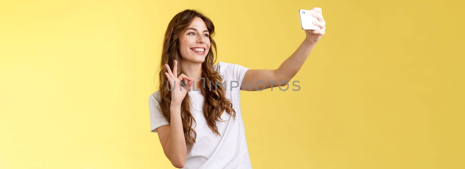 Stylish carefree urban confident curly-haired girl recording video blog followers extend arm hold smartphone taking selfie front camera show okay ok approval gesture yellow background. Lifestyle.