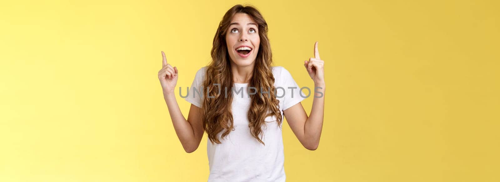 Happy impressed attractive girl long curly haircut drop jaw amused smiling surprised delighted look pointing up index fingers stare admiration fascinated awesome promo offer yellow background.Lifestyle.