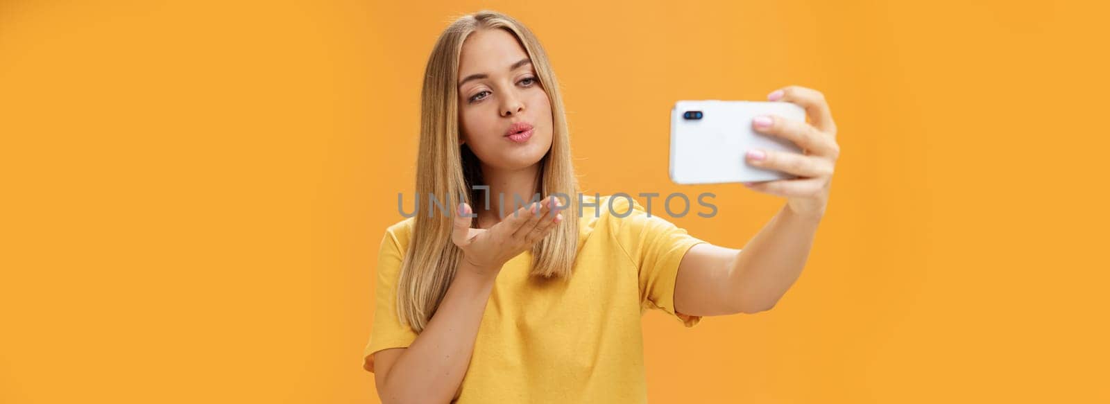 Stylish glamourous female fashion blogger ending recording video via smartphone by sending air kiss at camera, taking selfie with sensual and confident gaze at screen posing over orange wall. Lifestyle, blogger, van life concept