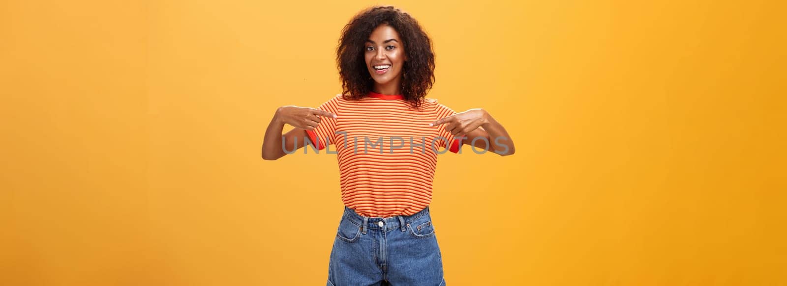 Hey pick me I your girl looking for. Portrait of charming friendly-looking ambitious dark-skinned female with afro hairstyle pointing at chest proudly and joyful posing against orange background.