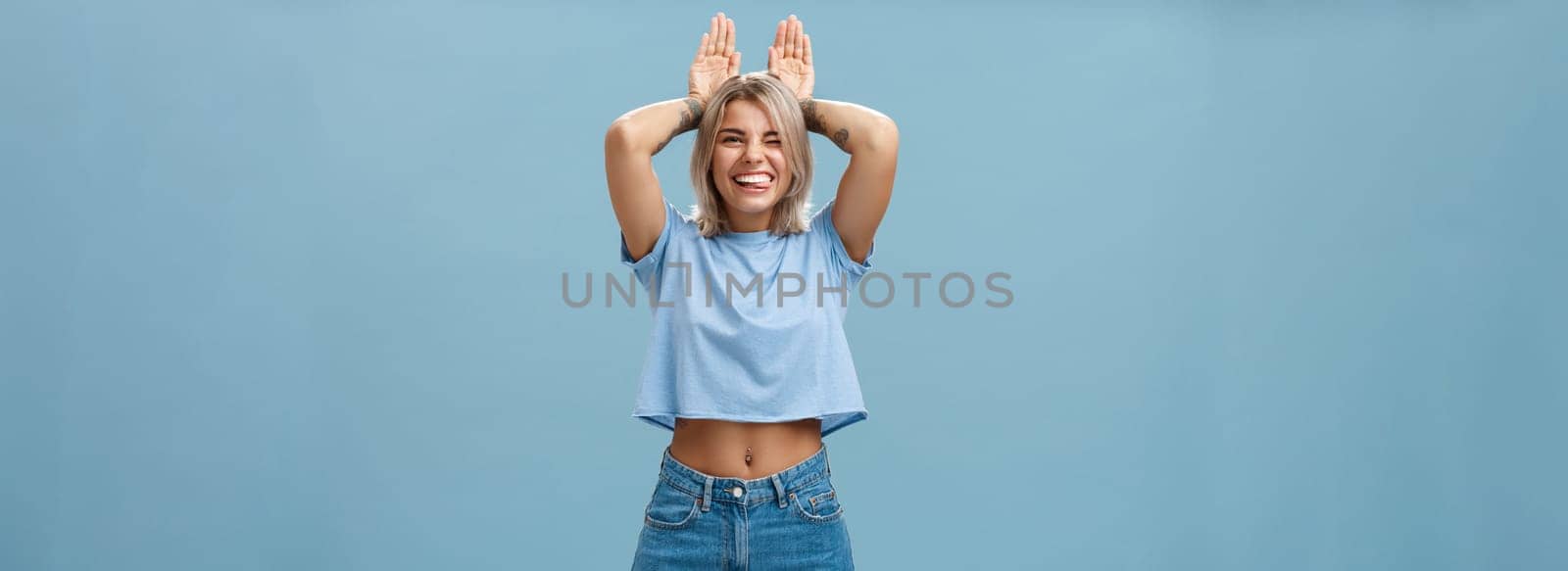 Time to relax. Portrait of happy playful cute blonde girl in trendy t-shirt with pierced belly and tattoo smiling joyfully holding palms on head like bunny ears having fun over blue background by Benzoix