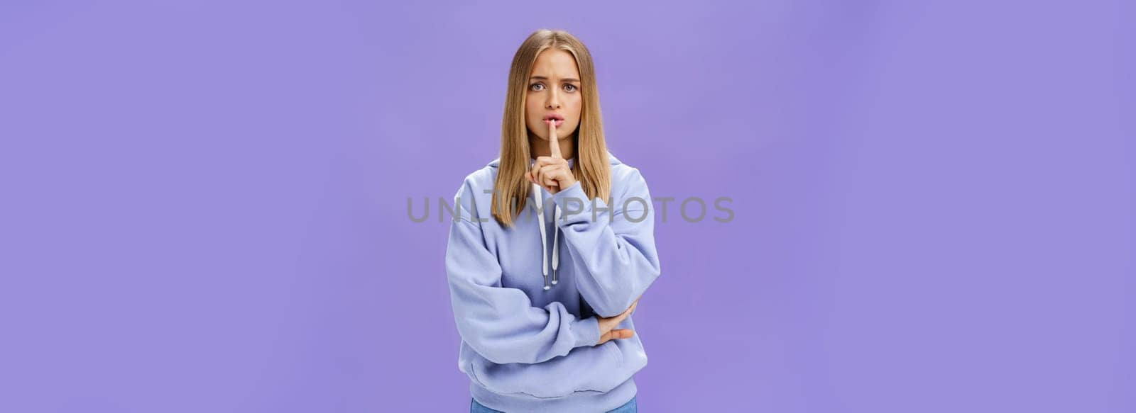 Girl concerned someone know secret making shush gesture with index finger over mouth beggin not tell anyone with worried serious look staying shh crossing one arm against chest timid and insecure by Benzoix