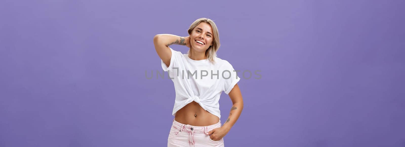 Indoor shot of carefree relaxed and joyful attractive athletic blonde woman in trendy summer outfit touching back of neck and smiling joyfully as if being in awkward situation over purple wall by Benzoix
