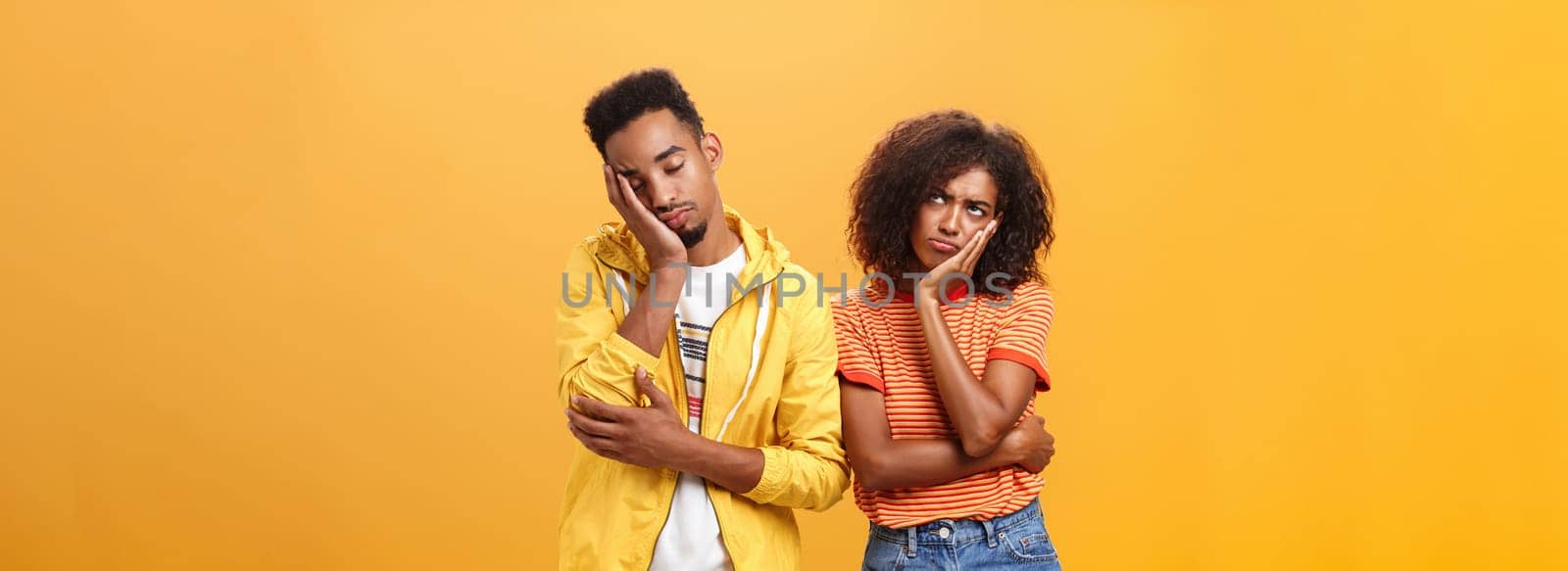 Girl feeling displeased and offended on boyfriend who fell asleep during date pursing lips frowning looking up while boyfriend leaning head on face with closed eyes and tired look over orange wall. Lifestyle.