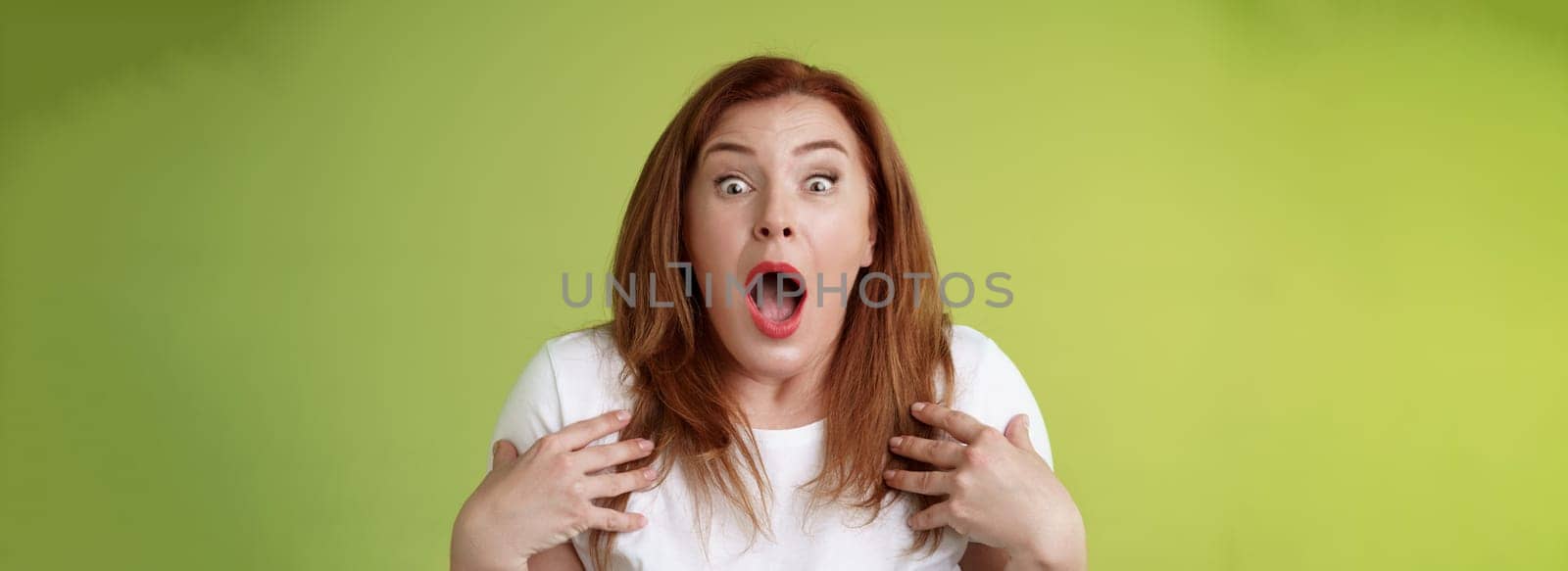 Shocked panicking redhead middle-aged woman gasping drop jaw open mouth stare camera freak-out anxious pointing herself impressed terrified frustrated nervously react green background.