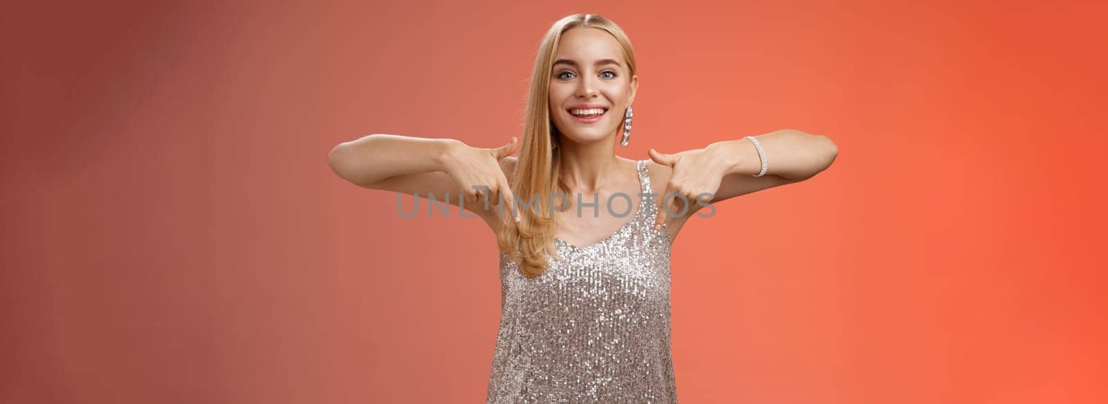 Lifestyle. Attractive glamour blond woman in silver glittering dress pointing down smiling excited showing awesome party place hang-out inviting try-out standing pleased red background enjoying perfect evening.