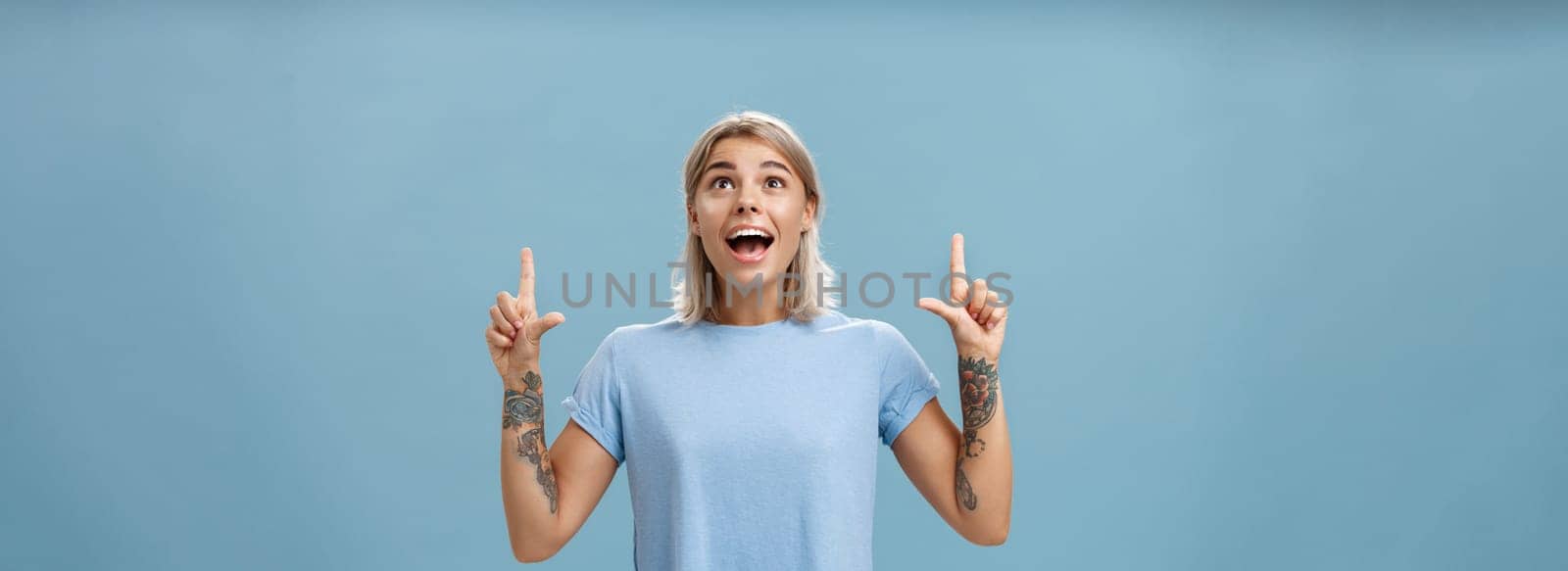 Studio shot of impressed speechless happy good-looking woman with tattoos on arms dropping jaw from amazement and joy gazing fascinated and pointing up standing over blue background by Benzoix