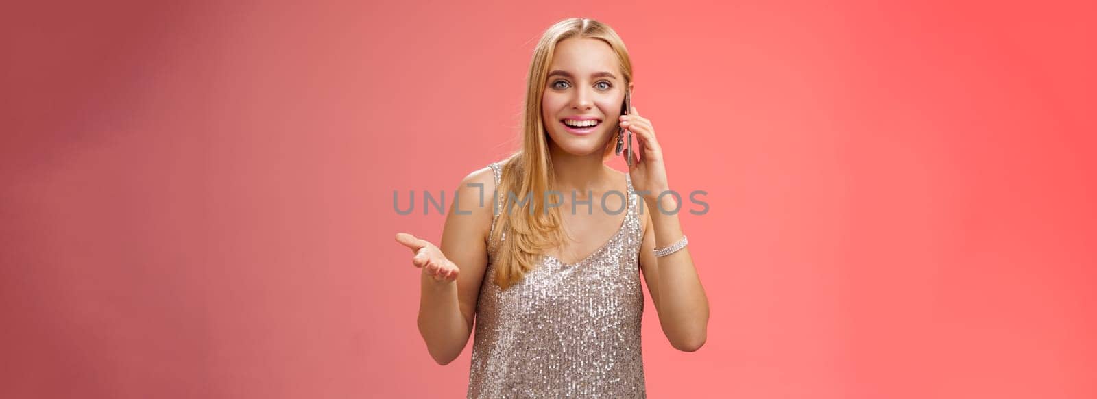 Joyful talkative outgoing attractive blond woman talking friend smartphone gesturing amused smiling broadly retelling fresh rumors after party wearing silver stylish dress, red background by Benzoix