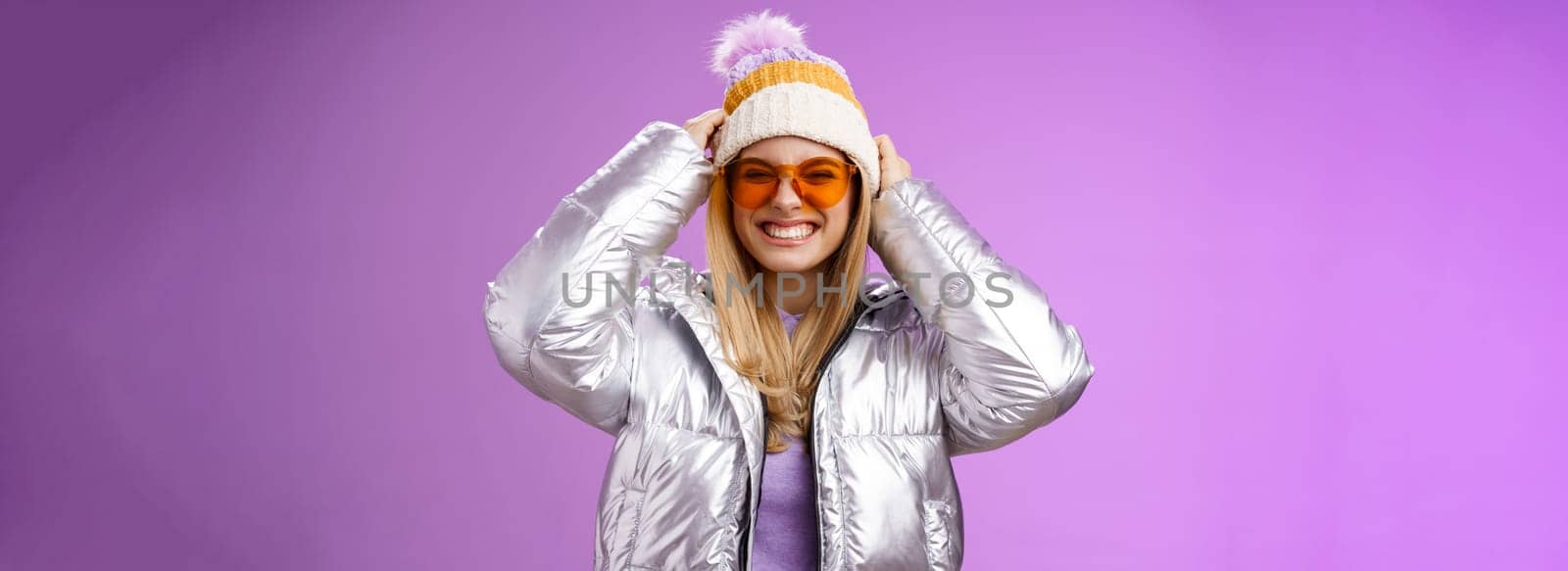Energized daring sassy young attractive woman having fun friends winter trip learn snowboarding smiling cheeky enjoying vacation put-on hat wearing silver warm jacket sunglasses, purple background.