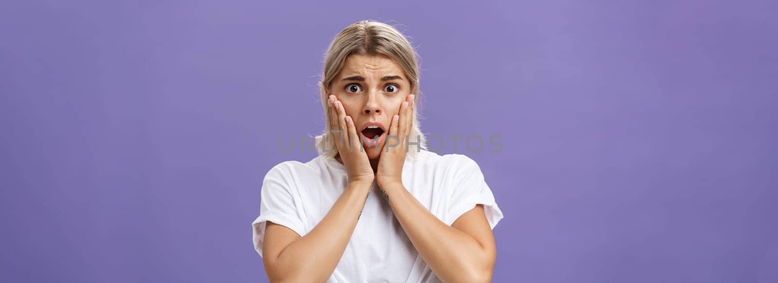 Shocked and worried silly european woman expressing empathy feeling sorry for poor animal in danger gasping opening mouth holding hands on face frowning feeling troubled over purple wall by Benzoix