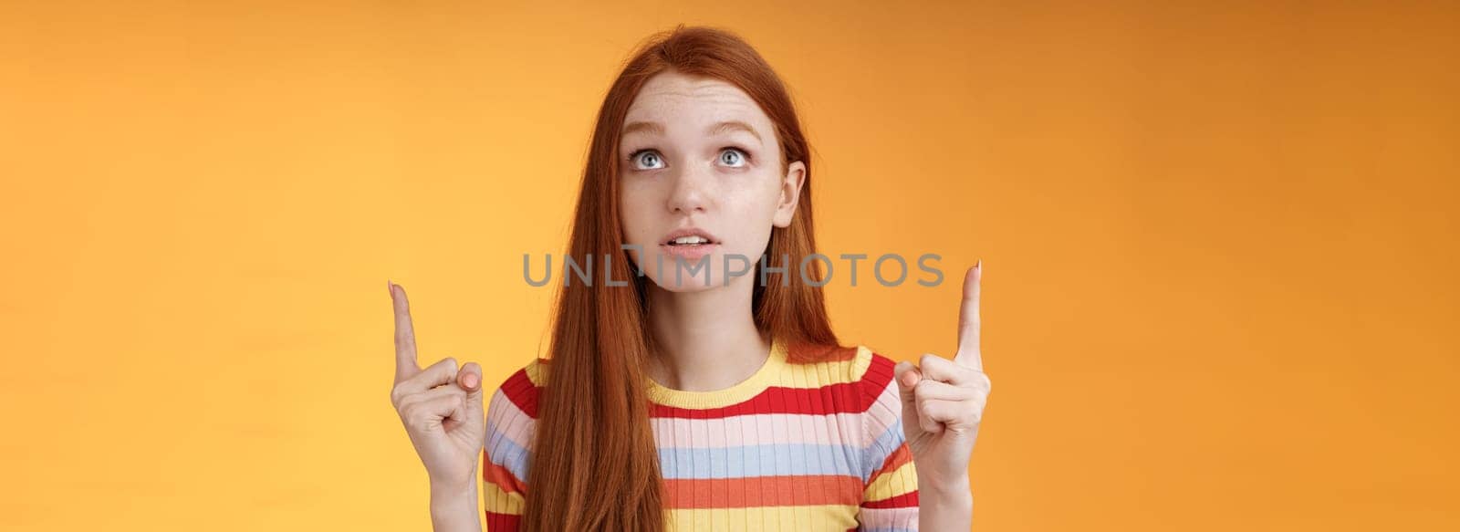 Stunned thrilled young redhead woman peer focused pointing up index fingers upwards look concentrated excited hold breath amused performance standing orange background intrigued and curious by Benzoix