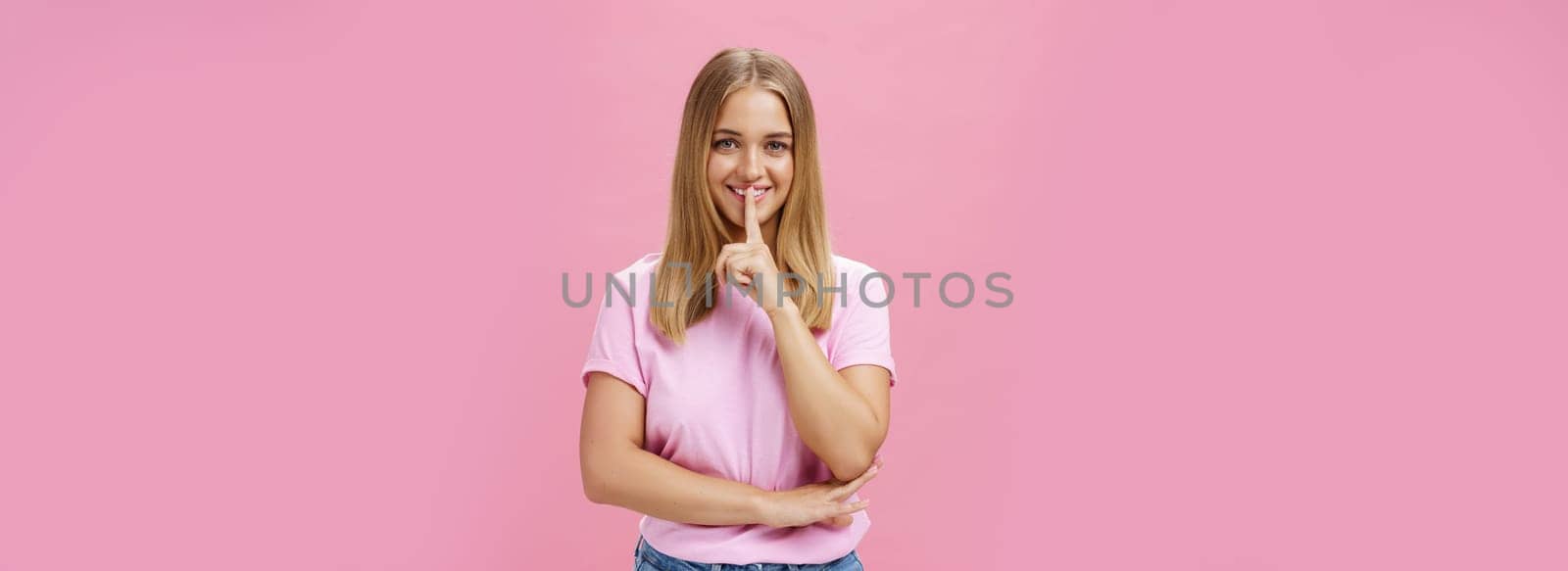 Shh keep it secret. Portrait of charismatic cheerful chubby girl with tanned skin and fair hair showing shush gesture with index finger over mouth smiling hiding surprise posing over pink background.