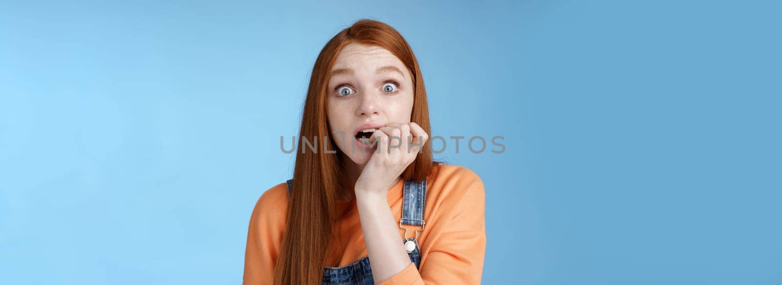 Scared unconfident anxious young trembling redhead girl wide eyes staring intense emotional biting fingernails, fan worry favorite character tv series dies standing nervously blue background.