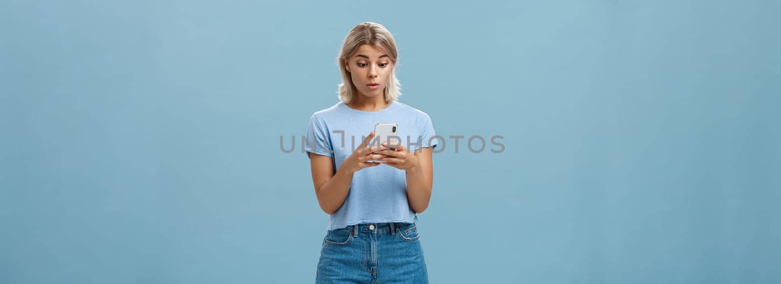 Indoor shot of surprised young caucasian girl receiving unexpected invitation via smartphone reading strange message in cellphone staring stunned and confused at screen over blue background by Benzoix