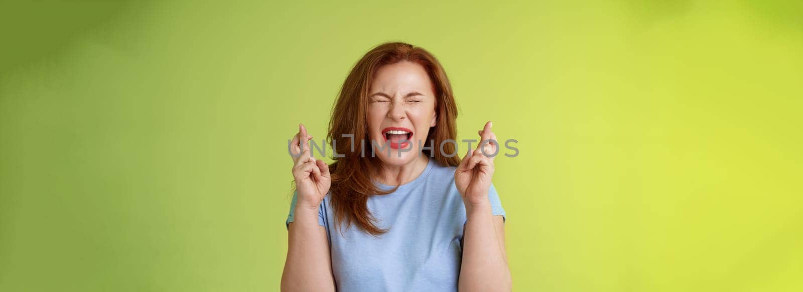 Woman wants win badly. Enthusiastic lucky redhead middle-aged 50s female pleading implore god make dream come true cross fingers good luck wishing closed eyes open mouth excitement green background.