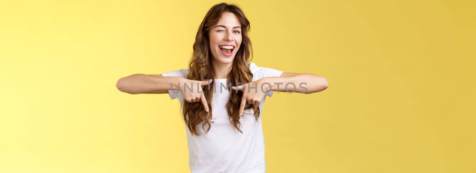 Hey visit my link would not regret. Lively cheeky enthusiastic woman curly long haircut wink sassy smiling broadly show you perfect copy space pointing down bottom promo yellow background by Benzoix