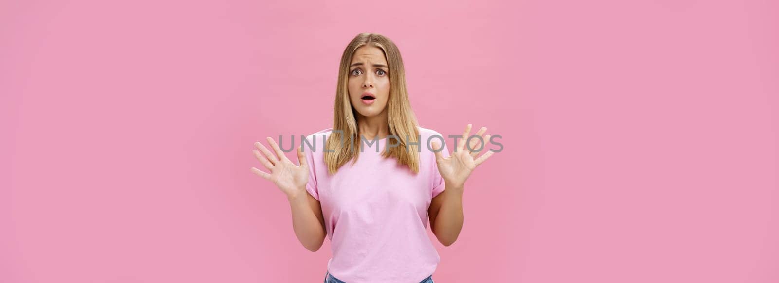 Woman looking nervous explaining with panicking gestures she not involved frowning opening mouth and gasping feeling concerned and worried waving hands over chest posing against pink wall by Benzoix