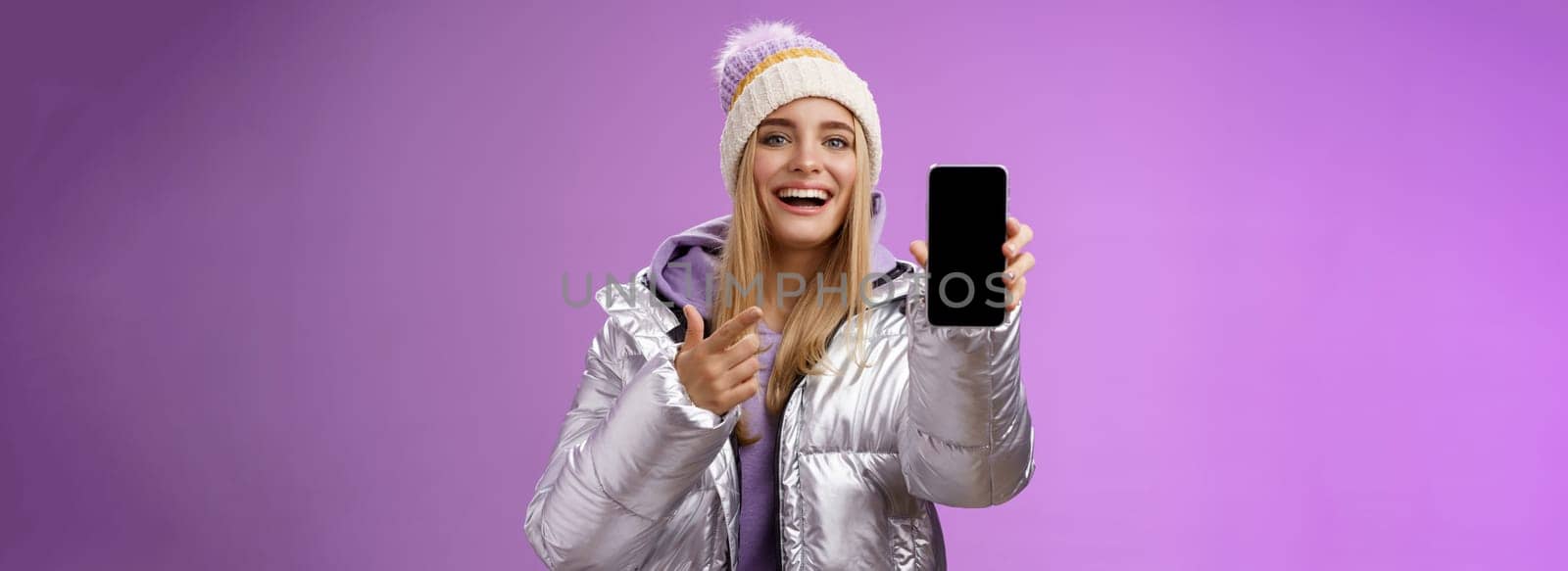 Satisfied amused good-looking blond girl suggest take look smartphone display smiling happily pointing mobile phone delighted talking about awesome new app features, standing purple background.