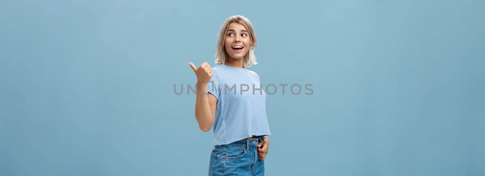 Charming sociable and friendly stylish female with blond hair turning right to show way or while discussing something talking with relaxed carefree look posing over blue background in trendy outfit by Benzoix