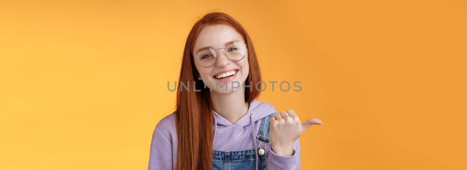 Cute helpful friendly-looking joyful european redhead woman show thumb left smiling delighted laughing pointing where find awesome store telling about interesting new promo offer, orange background.