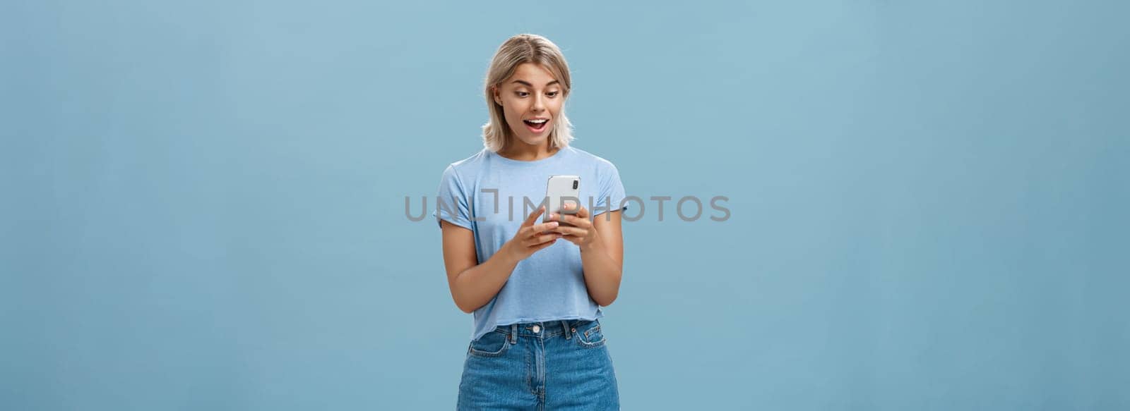 Entertained charming young european female with blond hair in casual outfit holding smartphone being satisfied with great camera and photos gasping and gazing amazed at screen over blue wall. Technology concept