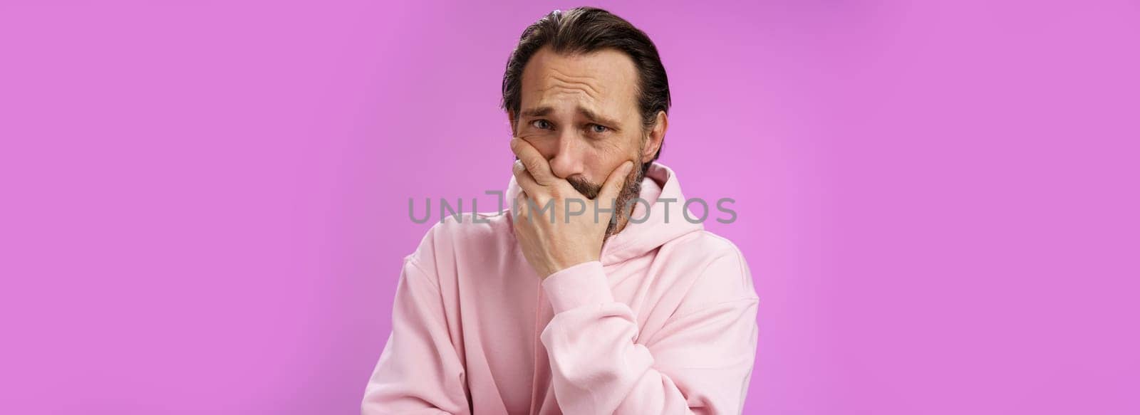 Shocked upset handsome mature bearded man hear terrible news griefing suffer sorrow express empathy gasping hold hand mouth reacting lose cringing crying cannot control emotions, purple background.