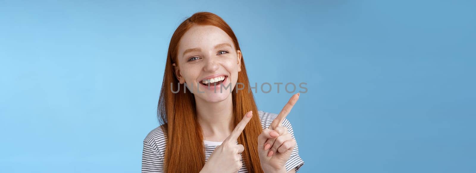 Happy carefree smiling redhead caucasian girl ginger straight hairstyle pointing upper left corner laughing joyfully amused cool promo giving recommendation try new product, standing blue background.