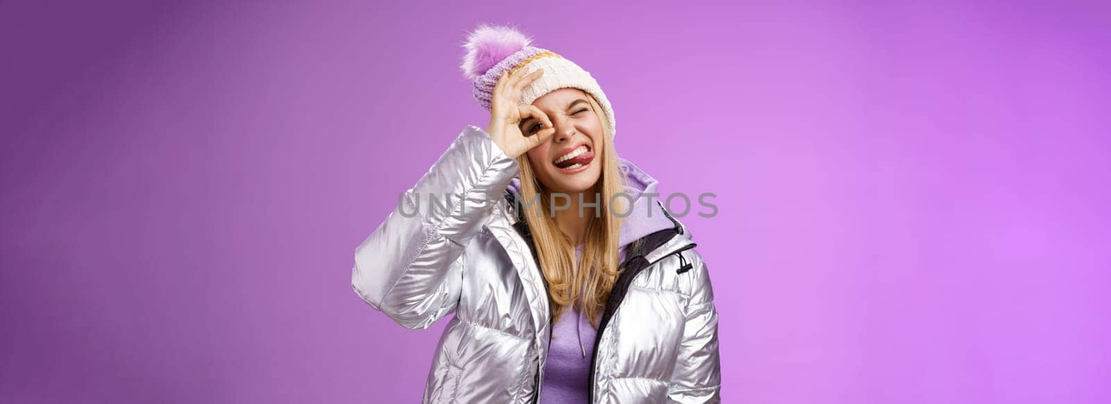 Friendly joyful caucasian blond girl in hat warm stylish shiny silver jacket tilt head happily show tongue smiling broadly enjoying awesome ski resort vacation travelling winter, purple background.