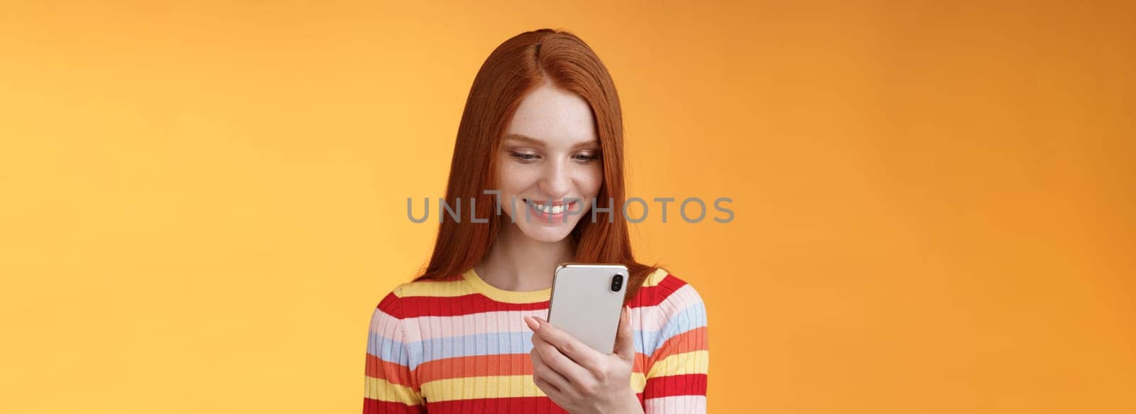 Charming modern redhead girl college student checking message box holding smartphone look happy smiling delighted cellphone display receive hundred likes photo post online, orange background.