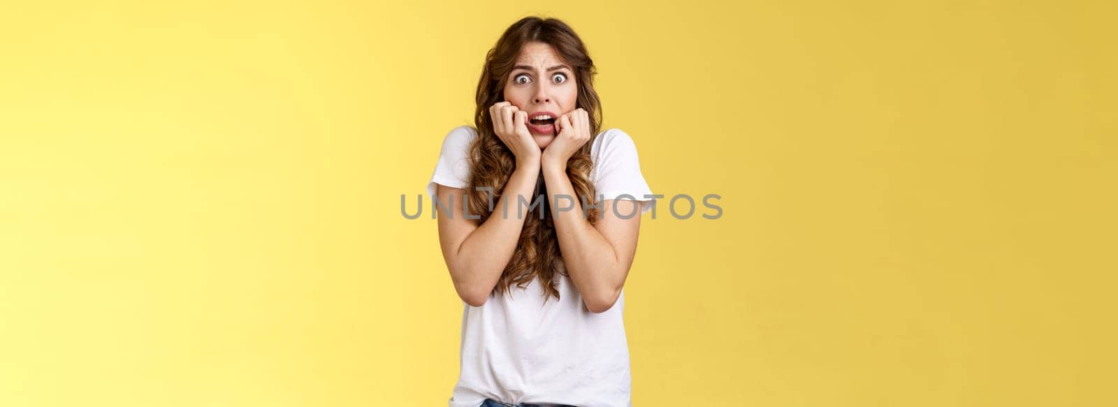 Timid insecure intense gasping young scared attractive woman sighing shocked frightened hold hands face biting nails terrified express fear standing shook yellow background stare camera by Benzoix