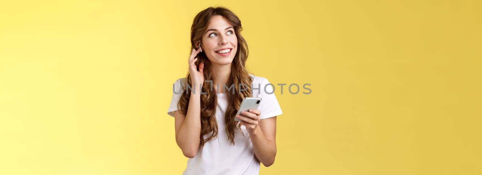 Dreamy happy cheerful curly-haired girl look around contemplate beautiful summer weather listening music touch wireless earbud calling friend talking via earphones hold smartphone yellow background. Lifestyle.