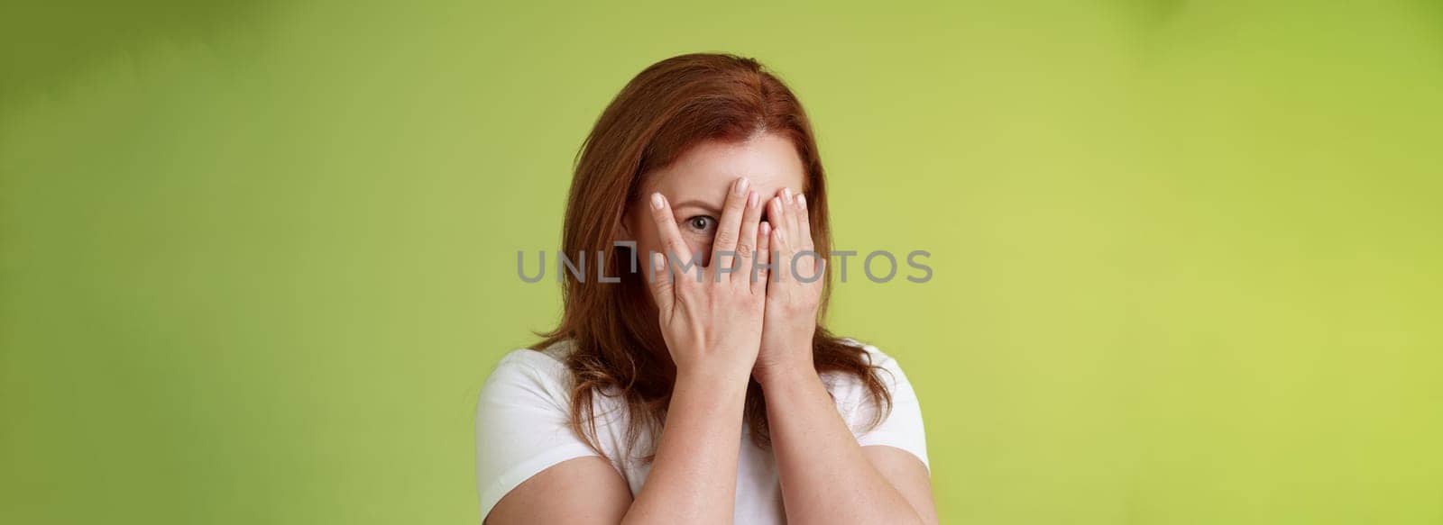 Not peeking. Playful charismatic middle-aged ginger redhead woman close face palms look through fingers one eye stare camera intrigued waiting surprise gift stand green background.