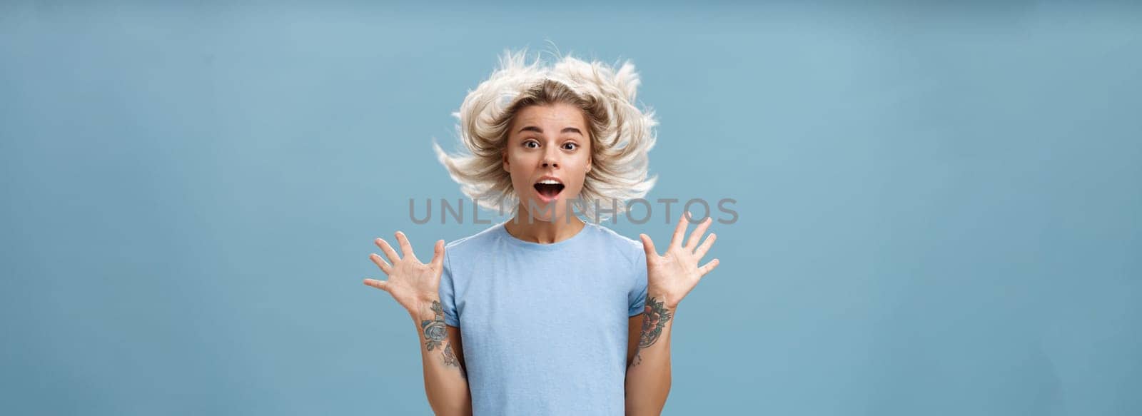 Waist-up shot of impressed surprised attractive and cute girl with blonde hair floating in air and tattooed arm jumping with spread raised palms and opened mouth over blue background by Benzoix