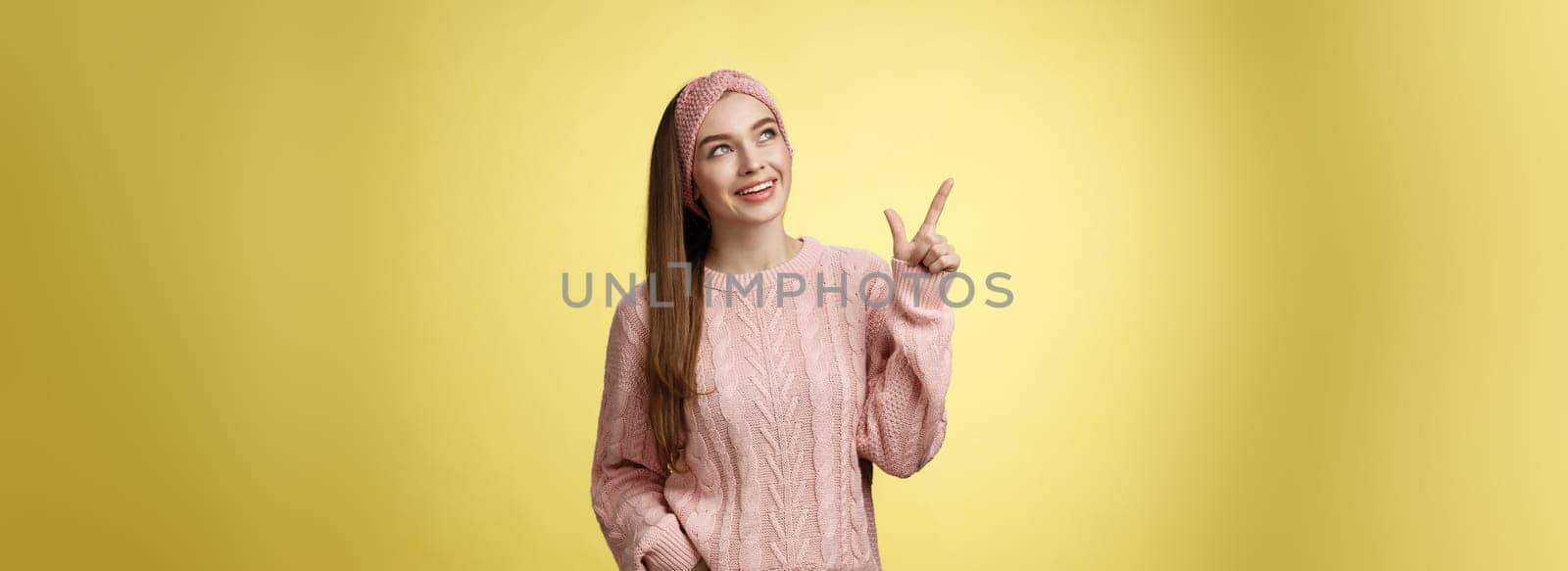 Portrait of amused interested cute 20s european girl wearing sweater, headband looking upper left corner pointing sideways smiling intrigued, ecstatic, captured by curious promo over yellow wall.