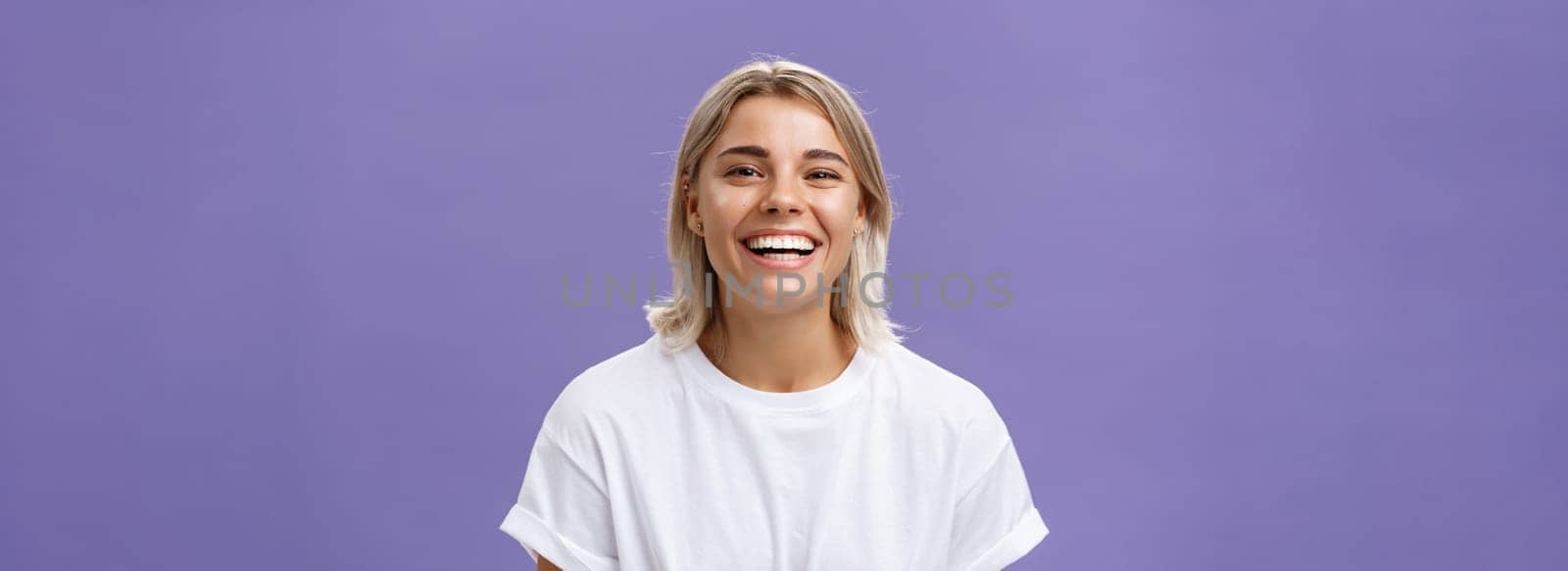 Close-up shot of joyful charming blonde female with delighted and pleased smile standing in white t-shirt over purple background spending time in awesome amusing company. Emotions concept