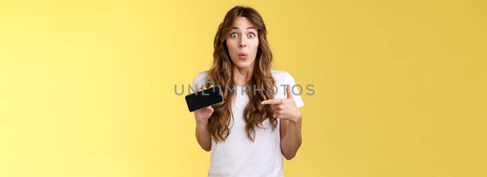 Surprised enthsuastic intrigued cute curly-haired caucasian girl folding lips amused curiously pointing smartphone screen stare camera ambushed astonished describe cool mobile phone app by Benzoix