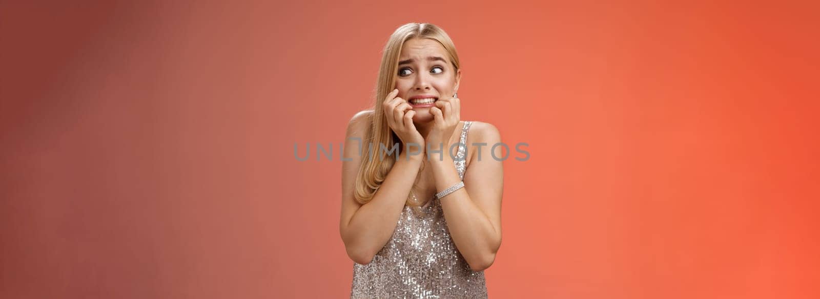 Lifestyle. Frightened insecure timid young blond woman afraid feel scared trembling fear stooping biting nails pop eyes right standing horrified red background wear silver evening dress terrified.