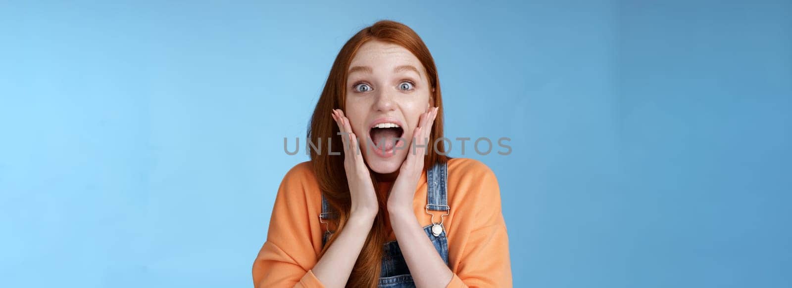 Excited thrilled young emotional enthusiasitc ginger girl teenage college student yelling amused smiling broadly receive positive good news look surprised camera touch face astonished blue background by Benzoix