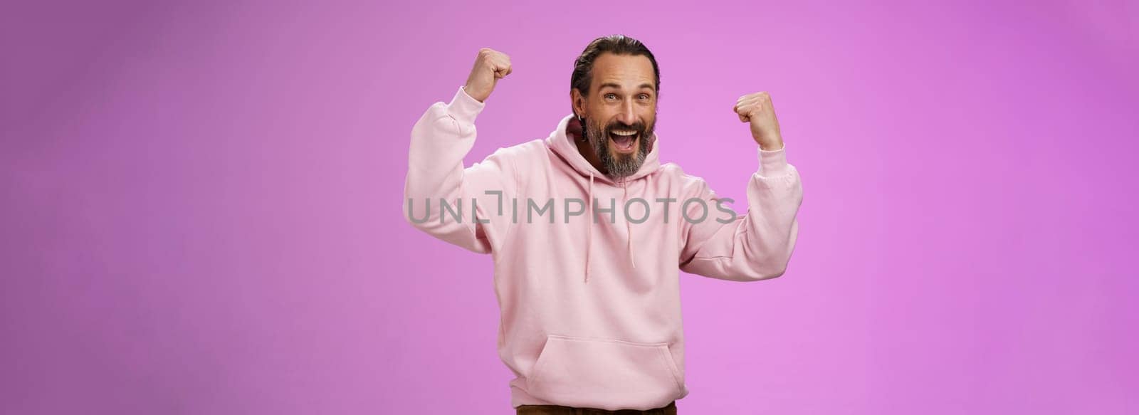 Yeah who cares age. Carefree delighted happy old man bearded in trendy pink hoodie raise fists joyfully triumphing having fun glad win celebrating success accomplish goal, posing purple background.