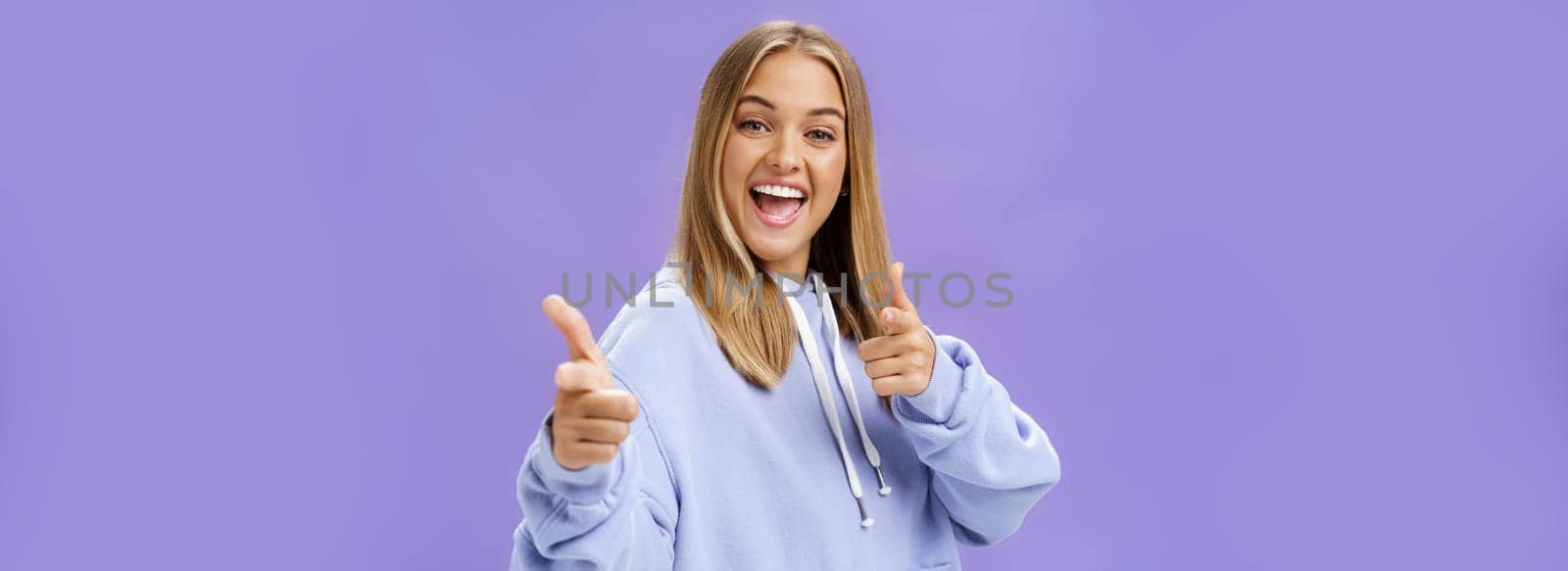 Enthusiastic and charismatic woman feeling joyful meeting friends checking out cool copy space pointing with finger guns at camera smiling broadly posing in stylish hoodie over purple wall. Copy space