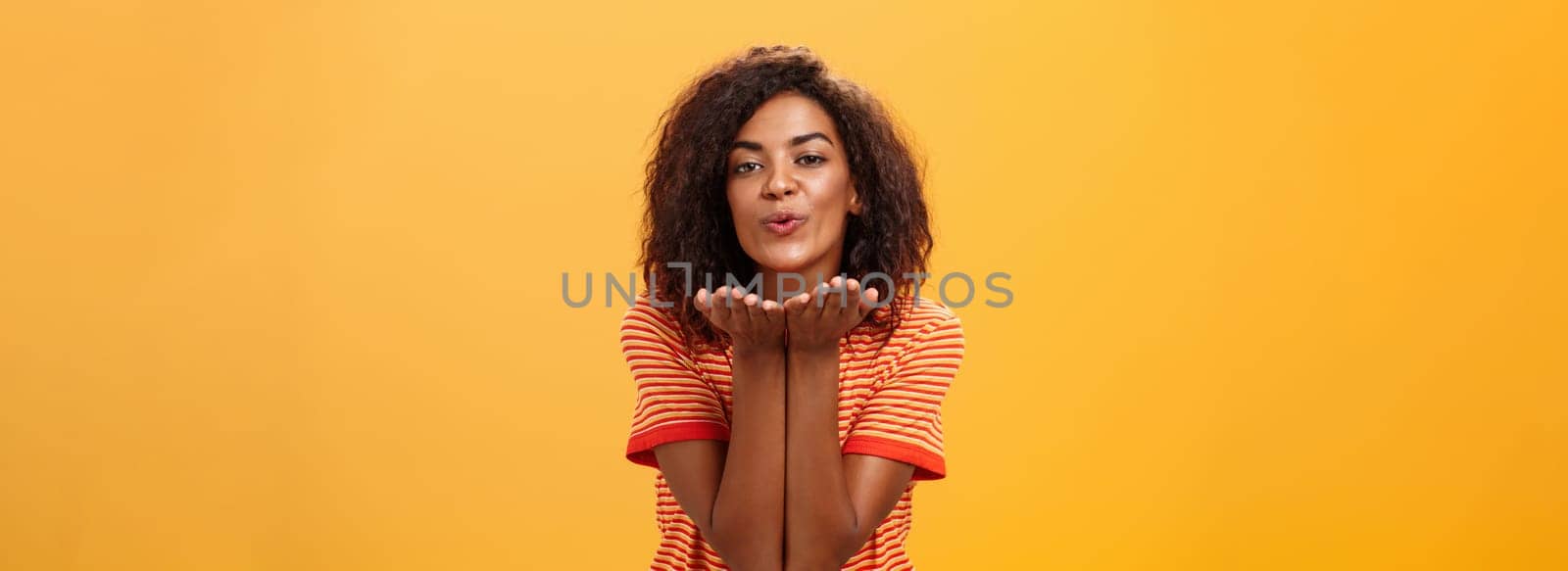 Sending passionate kiss to most loving person. Romantic attractive and stylish young african american girlfriend with curly hairstyle bending towards camera with slight smile, folded lips blowing mwah.