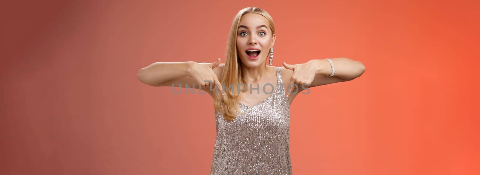 Impressed excited attractive glamour blond girl in silver glittering dress gasping thrilled pointing down glance camera fascinated check out fabulous awesome promo, standing amused red background by Benzoix