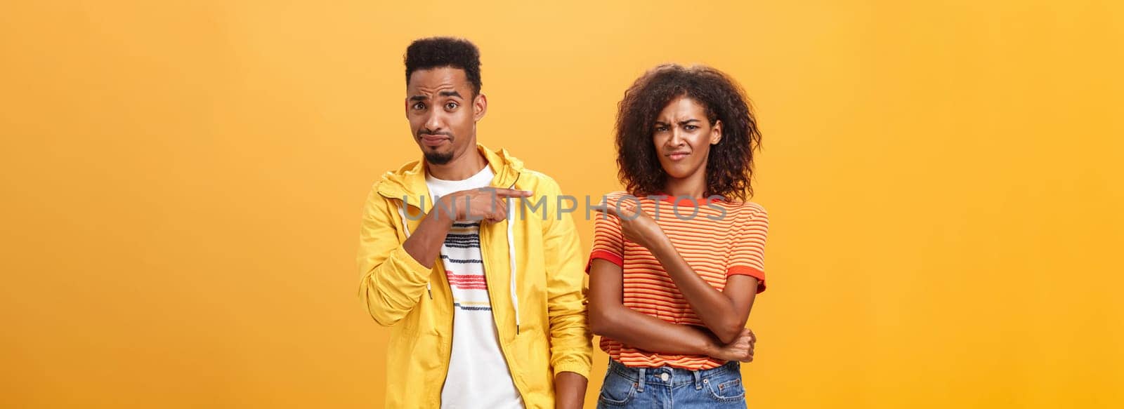 Friends getting shocked someone thought they are dating. Displeased mocking african american female frowning and grimacing from aversion while pointing at guy who indicating at girl over orange wall.