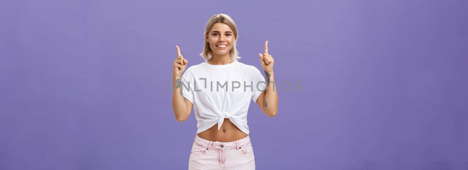 Time look upwards and move forward corporate ladder. Portrait of attractive ambitious and stylish young blonde woman with tattoos and pierced belly pointing up and smiling broadly over purple wall by Benzoix