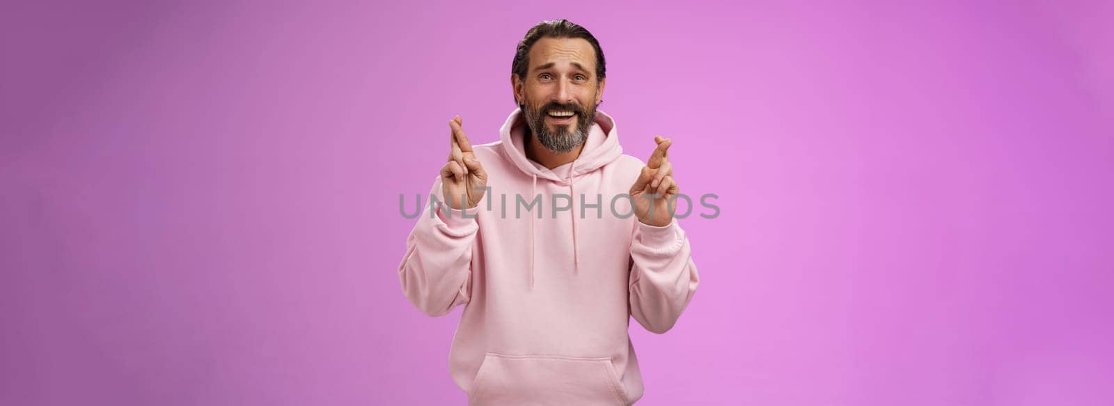 Hopeful lucky optimistic worried mature stylish bearded man grey hair in trendy hoodie cross fingers wish anticipating important results nervously standing purple background frowning anxious.