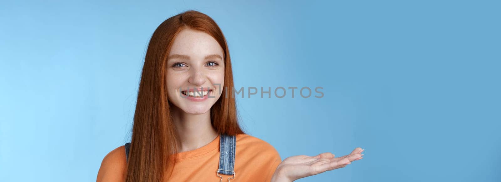 Proud good-looking confident redhead girl present awesome product hold object palm raise hand blank blue copy space smiling delighted recommend cool link, standing studio background helpful by Benzoix
