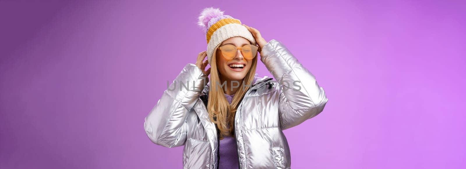 Carefree amused charming blond girlfriend having fun enjoying awesome sunny winter day ski resort vacation wearing sunglasses silver stylish jacket put on hat grinning joyfully, purple background by Benzoix